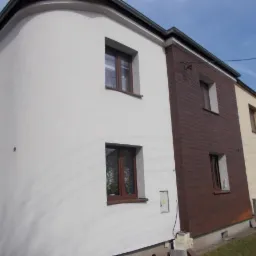 Dwukondygnacyjny dom z częściowo ocieploną i otynkowaną elewacją w kolorze białym, brązowe panele imitujące drewno na fragmencie ściany, brązowe ramy okienne, widoczne elementy instalacji...