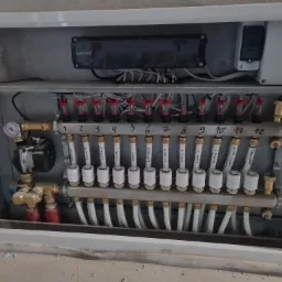 Rozdzielacz ogrzewania podłogowego z pompą obiegową, zaworami termostatycznymi i opisami stref grzewczych (jadalnia, salon, pokój, łazienka, kuchnia, garaż), zamontowany w szarej szafce...