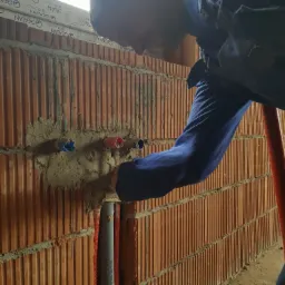 Instalacja rur wodnych w ścianie z pustaków ceramicznych; widoczne fragmenty niebieskich i czerwonych rur wystających ze ściany, oraz szara rura kanalizacyjna w czerwonej osłonie; hydraulik...