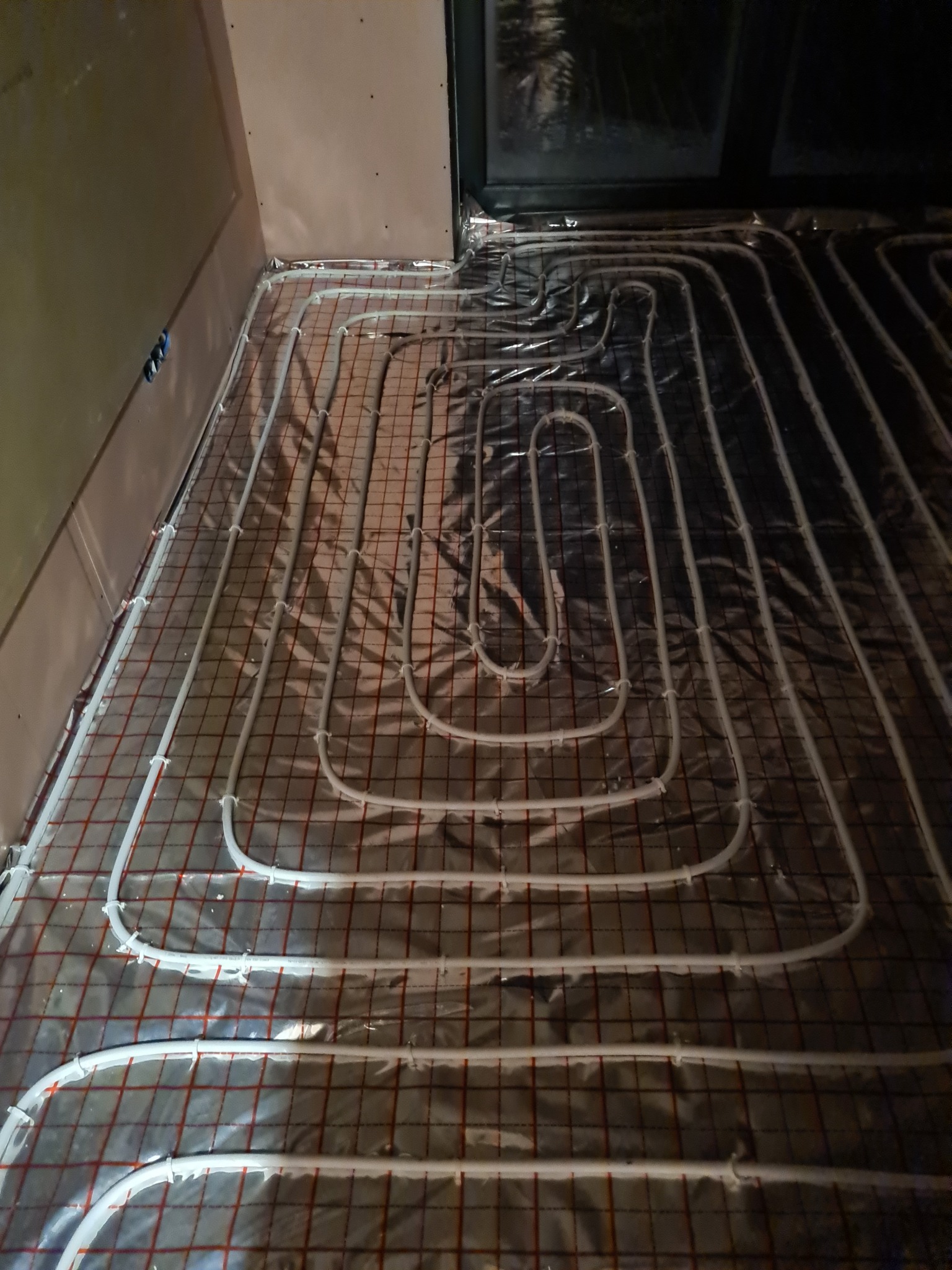 Rozłożone rury ogrzewania podłogowego na folii izolacyjnej z widoczną siatką, przygotowane do zalania wylewką.