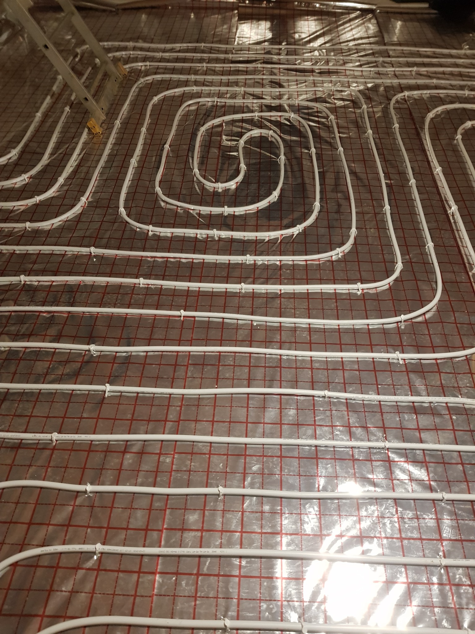 Białe rury ogrzewania podłogowego ułożone spiralnie na folii z czerwoną siatką, widoczny fragment drabiny w lewym górnym rogu.
