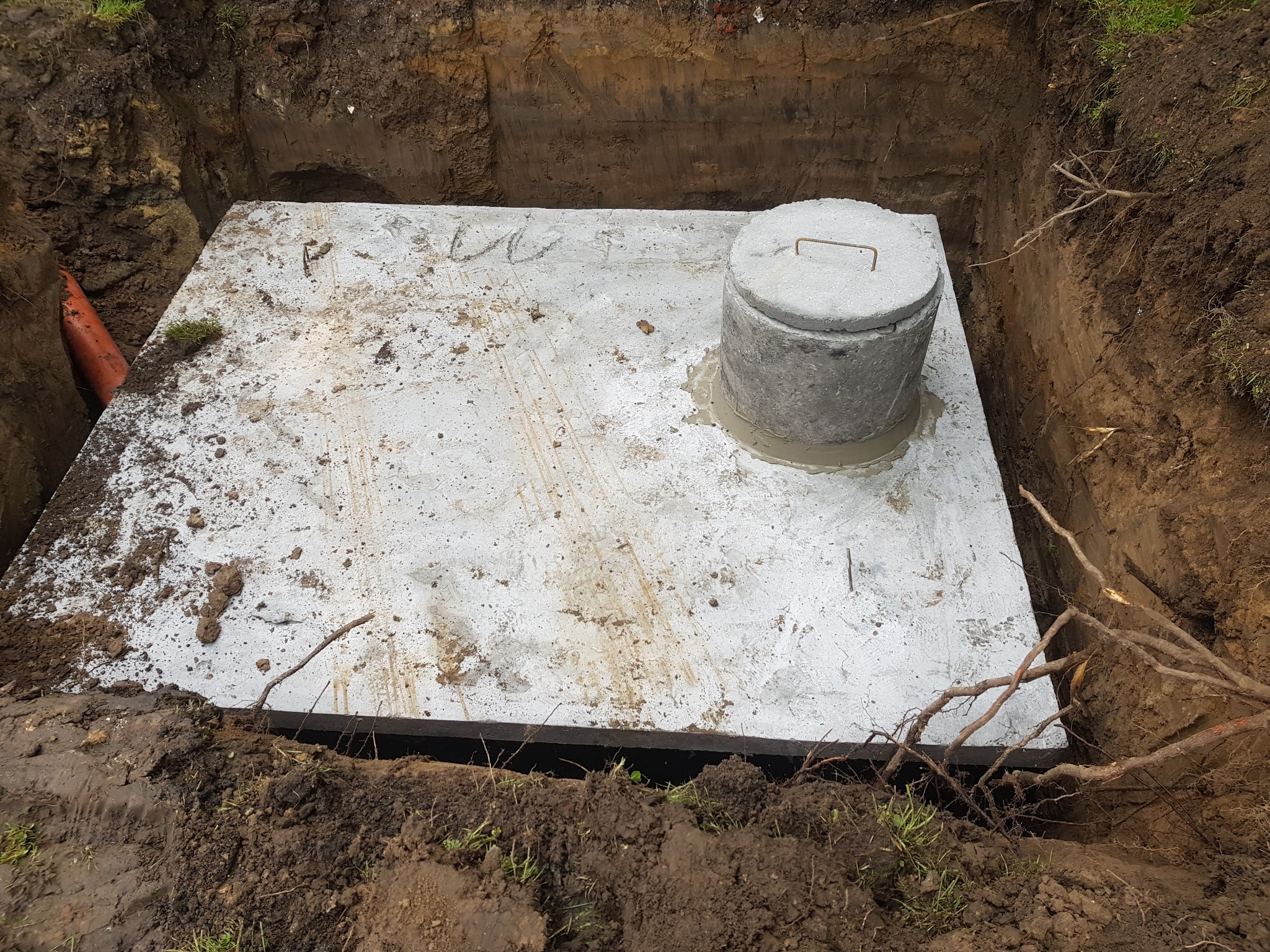 Betonowa płyta przykrywająca zbiornik, umieszczona w wykopie ziemnym. Widoczna pokrywa włazu z uchwytem oraz fragment pomarańczowej rury po lewej stronie.