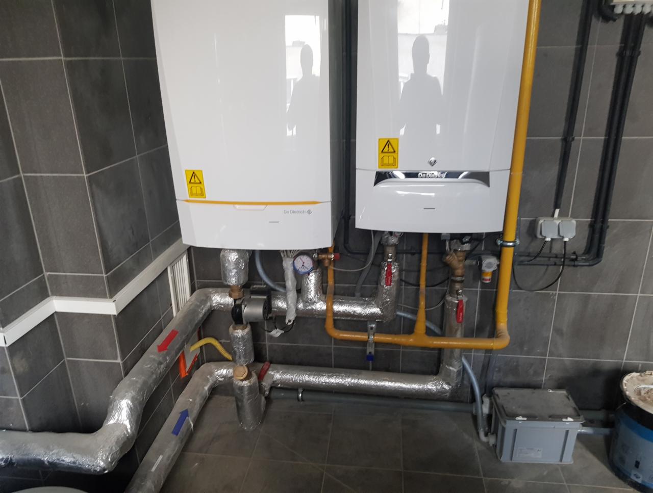 Dwa wiszące, białe piece gazowe marki De Dietrich z widocznymi rurami gazowymi w kolorze żółtym oraz rurami grzewczymi z izolacją, zamontowane na tle szarych, kwadratowych płytek.