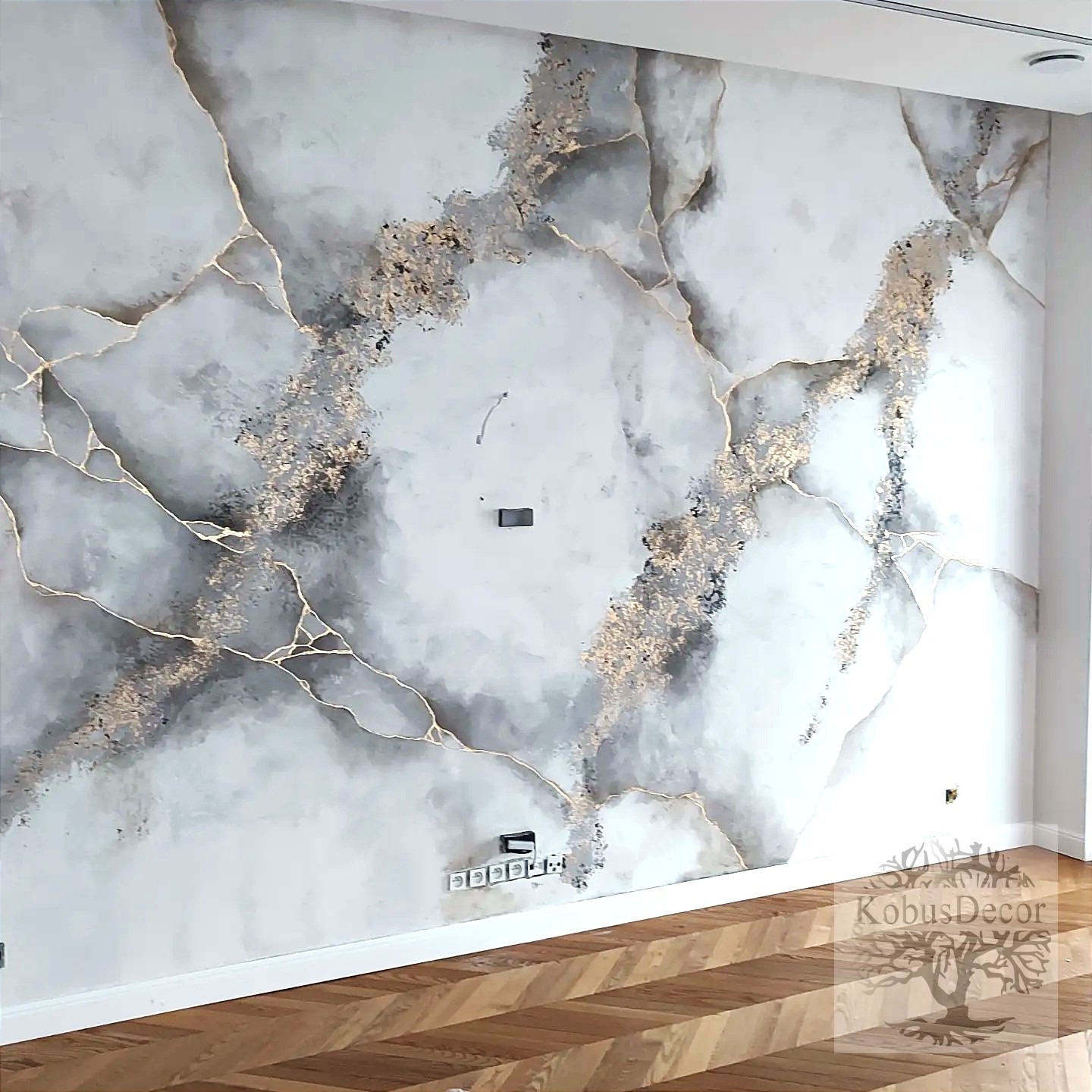 Ściana z dekoracyjną fakturą imitującą marmur z żyłkami złota, widoczne gniazdka elektryczne i drewniana podłoga ułożona w jodełkę.