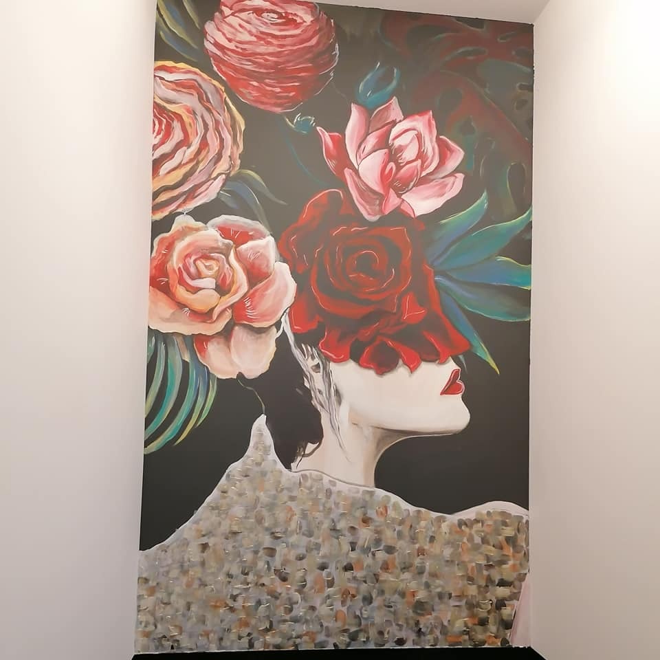 Artystyczny mural na ścianie przedstawiający profil kobiety z kwiatami zamiast oczu, w kolorach czerwieni, różu i zieleni na ciemnym tle.