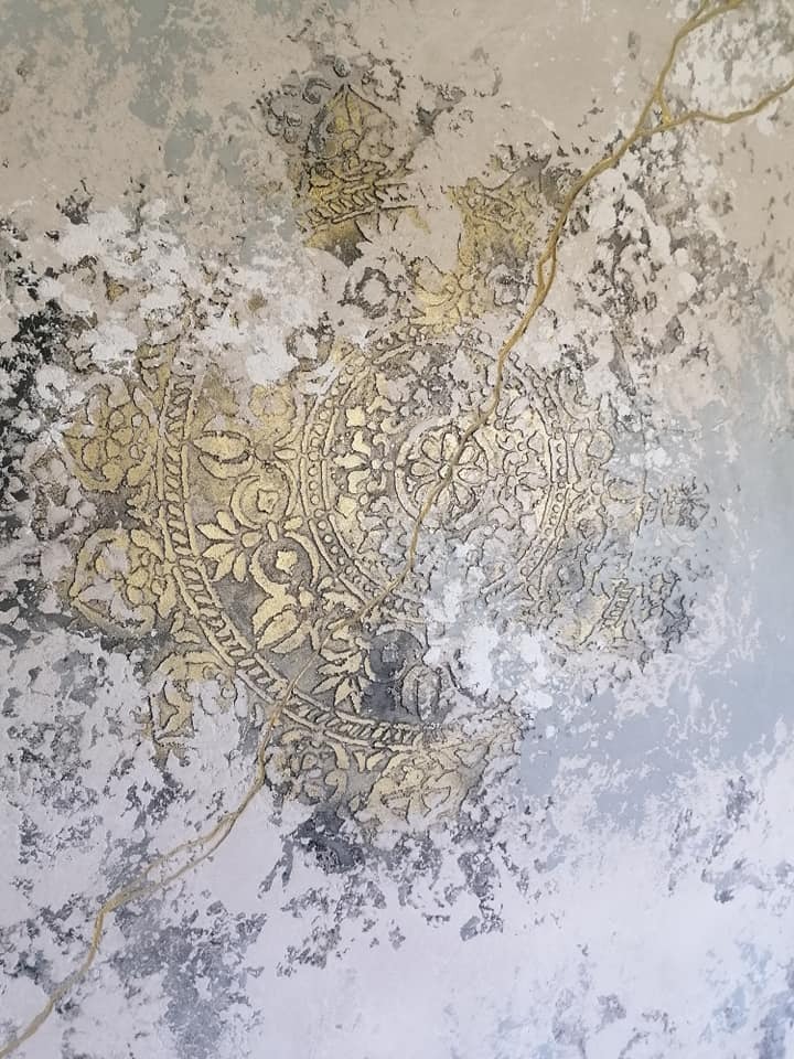 Dekoracyjna ściana z efektem stiuku w odcieniach szarości i złota, z wyraźnym motywem okrągłym o orientalnym wzorze, poprzecinana złotymi liniami imitującymi pęknięcia.