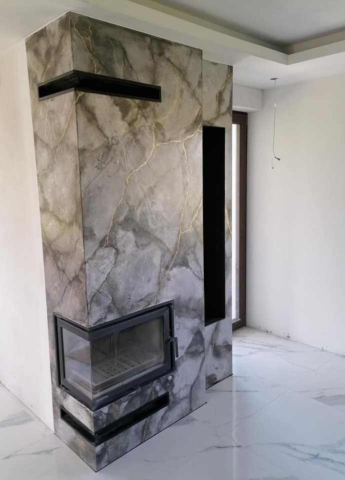 Obudowa kominka wykończona szarym stiukiem w marmurowy wzór z delikatnymi, złotymi żyłkami, czarne elementy dekoracyjne i rama wkładu kominkowego, biała marmurowa podłoga.