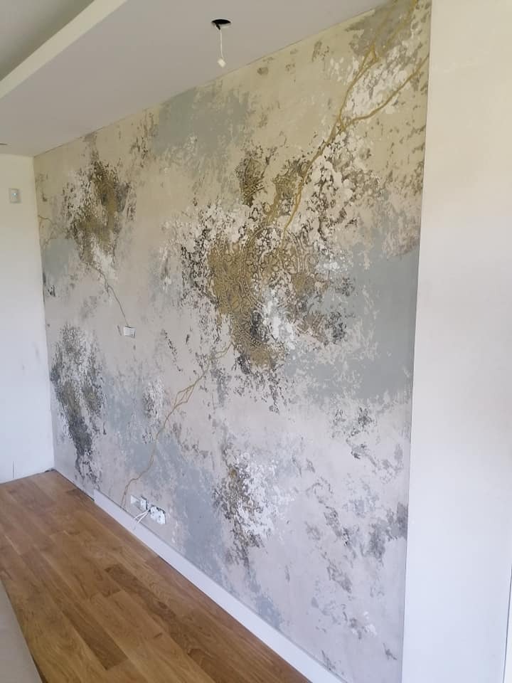 Ściana w pokoju z dekoracyjną fakturą imitującą marmur w odcieniach szarości, bieli i złota, z drewnianą podłogą. Widoczne gniazdka elektryczne przy podłodze i otwór w suficie po lampie.