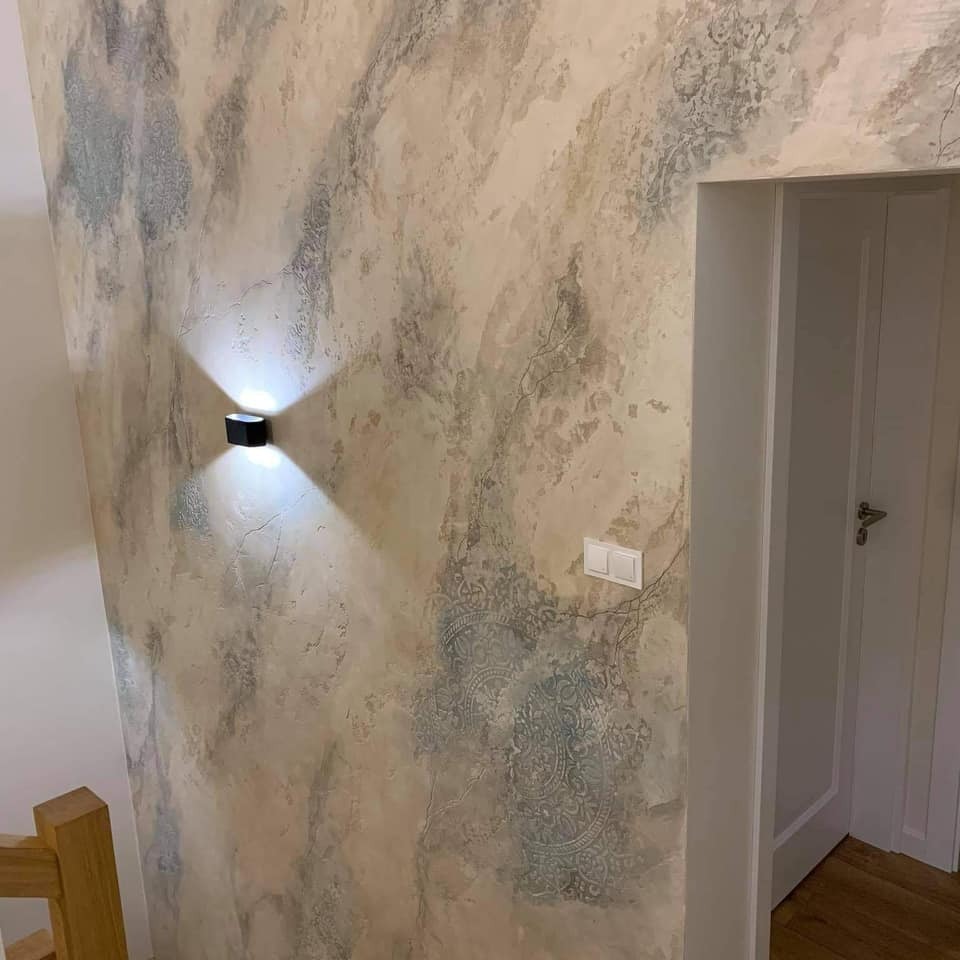 Ściana z dekoracyjną fakturą imitującą marmur i subtelnymi wzorami, oświetlona nowoczesnym kinkietem, obok białe drzwi z widocznym fragmentem podłogi.