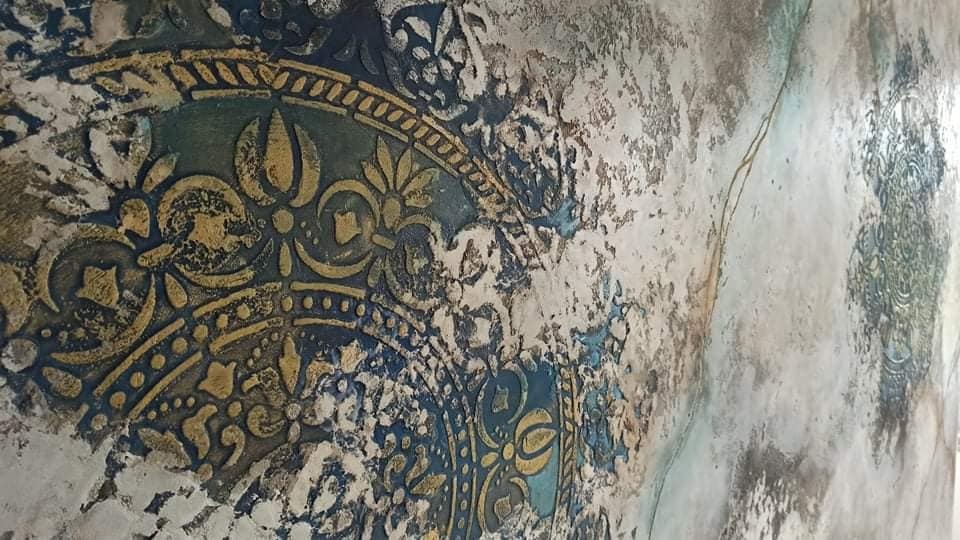 Fragment ściany z dekoracyjnym, wypukłym wzorem w odcieniach złota i błękitu, miejscami z efektem spękań i postarzenia.