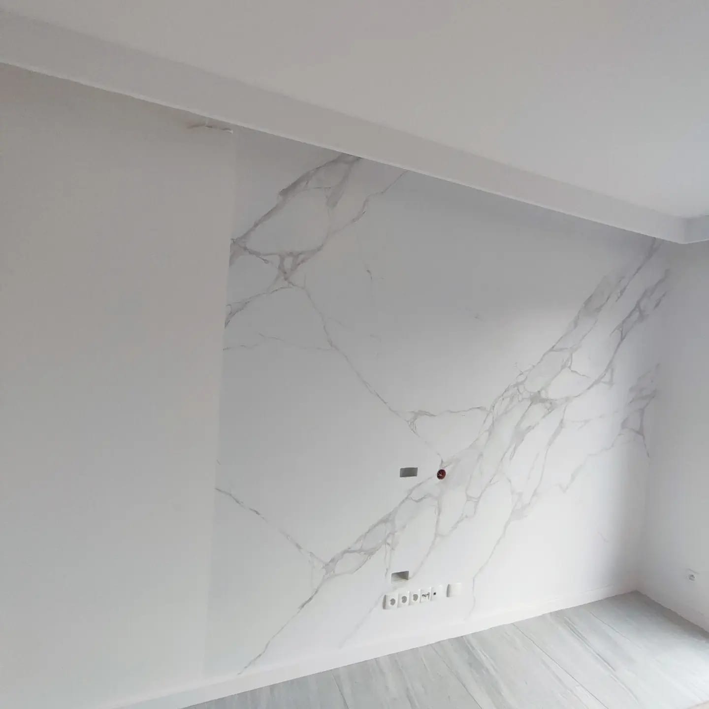 Ściana wykończona płytami imitującymi biały marmur z szarymi żyłami, widoczne gniazdka elektryczne i włączniki, podłoga w jasnych odcieniach szarości.