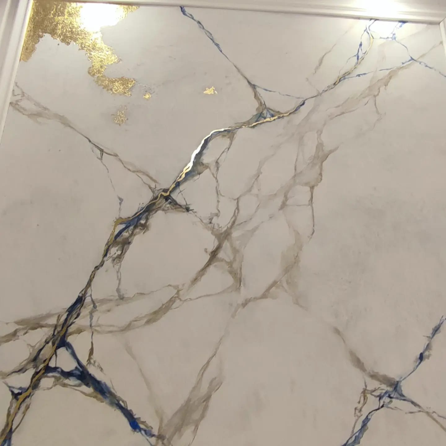 Ściana z efektem marmuru carrara z niebieskimi i złotymi żyłami oraz płatkami złota w górnym rogu, wykończona białą listwą przysufitową.