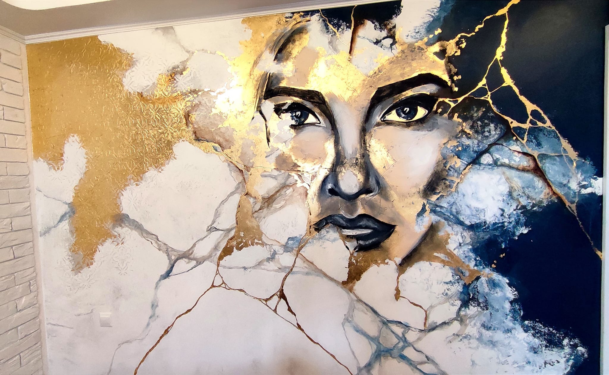 Abstrakcyjny portret kobiety z elementami złota na ścianie w jasnym pomieszczeniu, widoczny fragment ceglanej ściany.