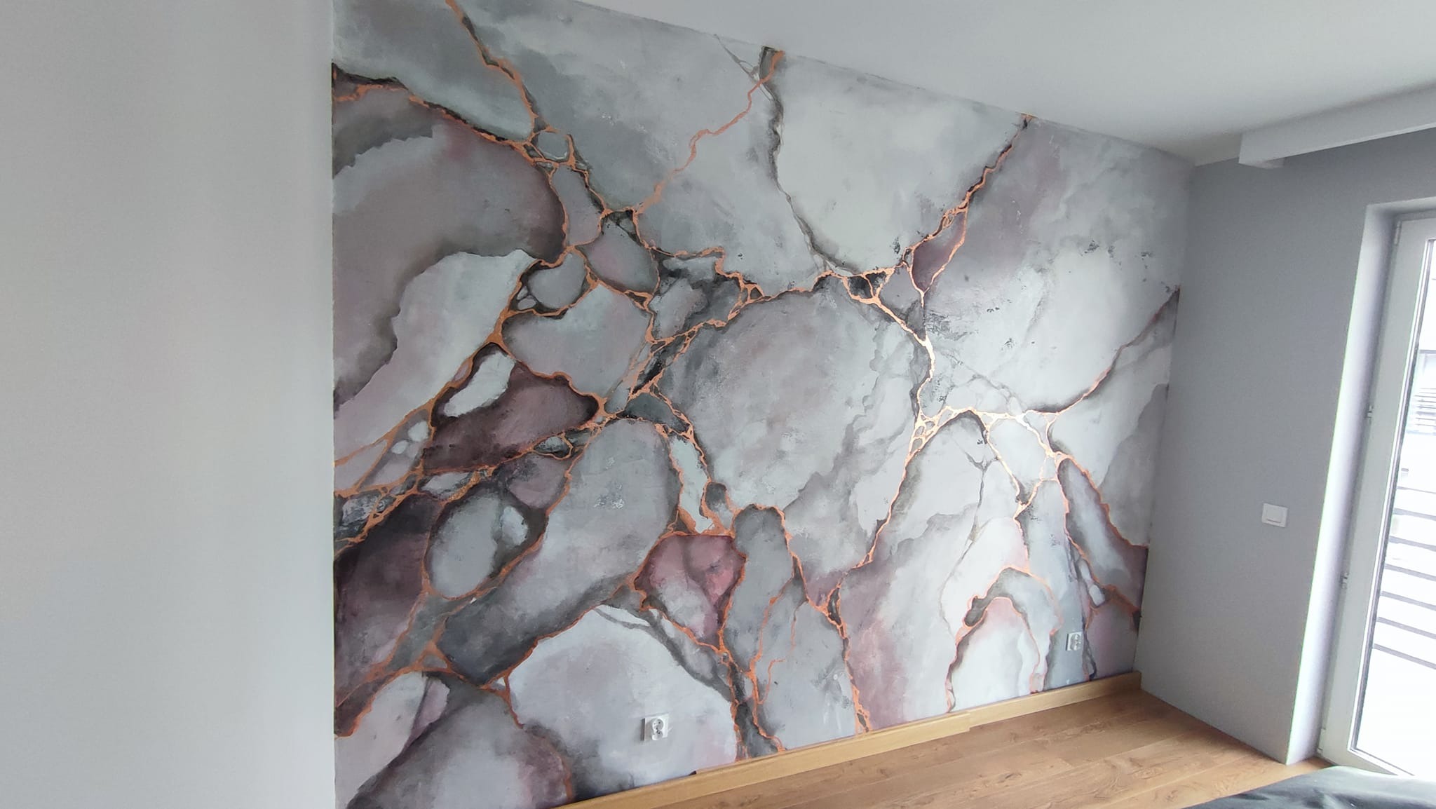 Ściana z dekoracyjną fakturą imitującą marmur w odcieniach szarości, różu i bieli, z żyłkami w kolorze miedzianym, widoczne gniazdko elektryczne.