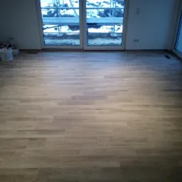 Ułożona podłoga z płytek imitujących drewno w nowym, pustym pokoju z dużymi oknami balkonowymi i widocznym rusztowaniem na zewnątrz.