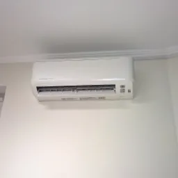 Biały klimatyzator Mitsubishi Inverter zamontowany na ścianie w jasnym pomieszczeniu, widoczny fragment okna i ozdobna listwa sufitowa.