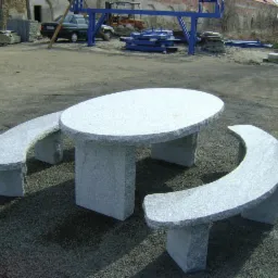 Komplet ogrodowy ze stołem i ławkami z jasnego granitu, wykonany w Strzegomiu, na tle placu z zaparkowanym samochodem i elementami konstrukcyjnymi.