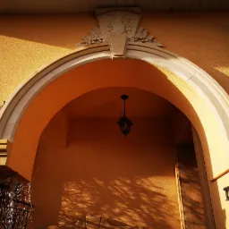 Wejście do budynku z łukiem z jasnego kamienia, zwieńczonym dekoracyjnym herbem, oświetlone wiszącą lampą w stylu retro, na tle pomarańczowej ściany z cieniami drzew.