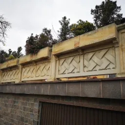 Długi, dekoracyjny mur z piaskowca z geometrycznym wzorem, zwieńczony donicami z roślinami, postawiony na murze z kamienia polnego, widok z dołu.