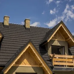 Dom z ciemną, nową dachówką i drewnianymi elementami wykończeniowymi, widoczne kominy z kamienia, błękitne niebo w tle.