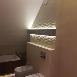 Nowoczesna łazienka na poddaszu ze ścianą dekoracyjną z podświetleniem LED, bidetem i toaletą. Uwagę przykuwa falista struktura ściany i ciepłe oświetlenie.