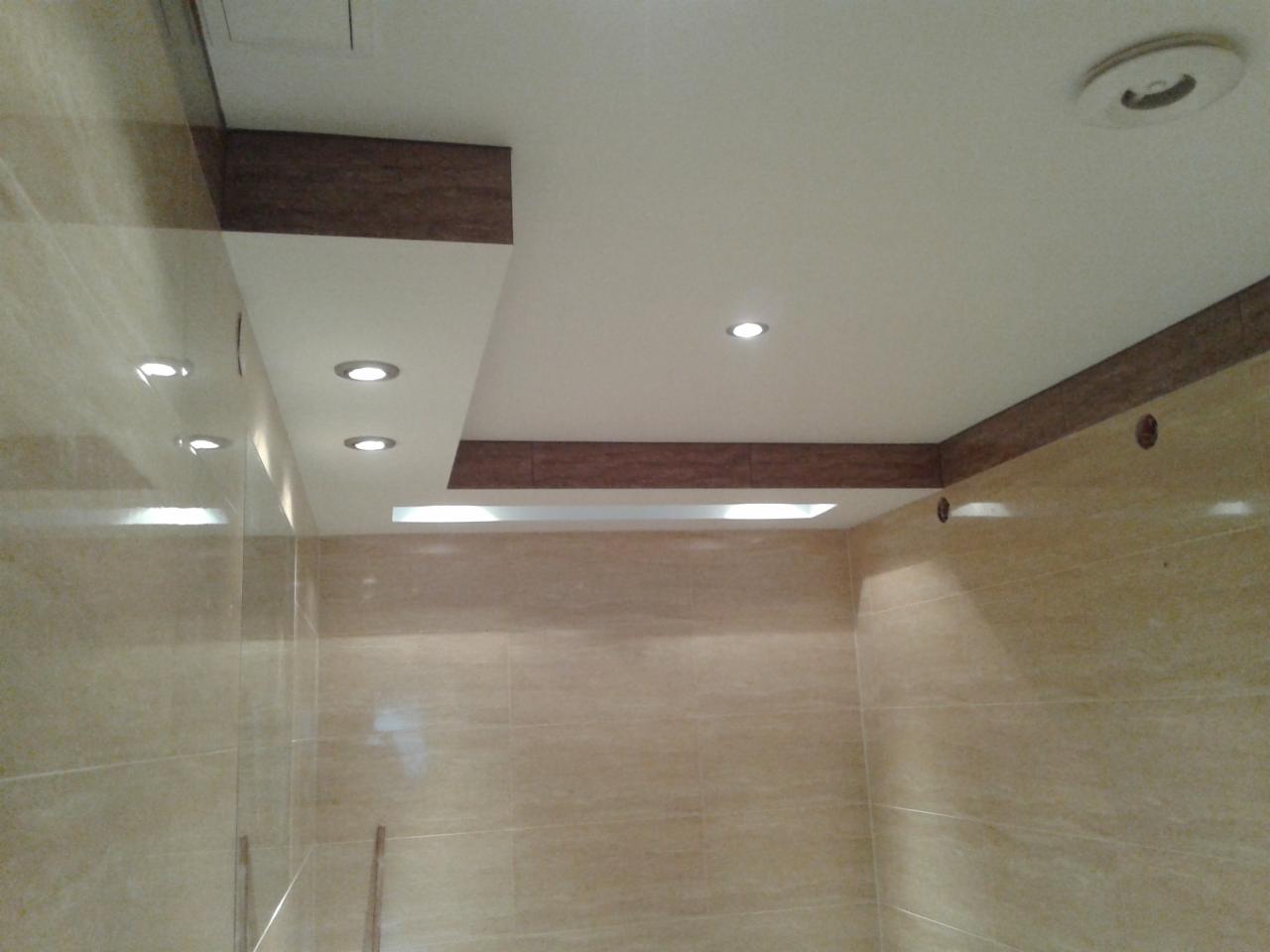 Beżowe płytki ścienne w łazience z podwieszanym sufitem z oświetleniem punktowym i drewnianymi akcentami. Widok na narożnik pomieszczenia.