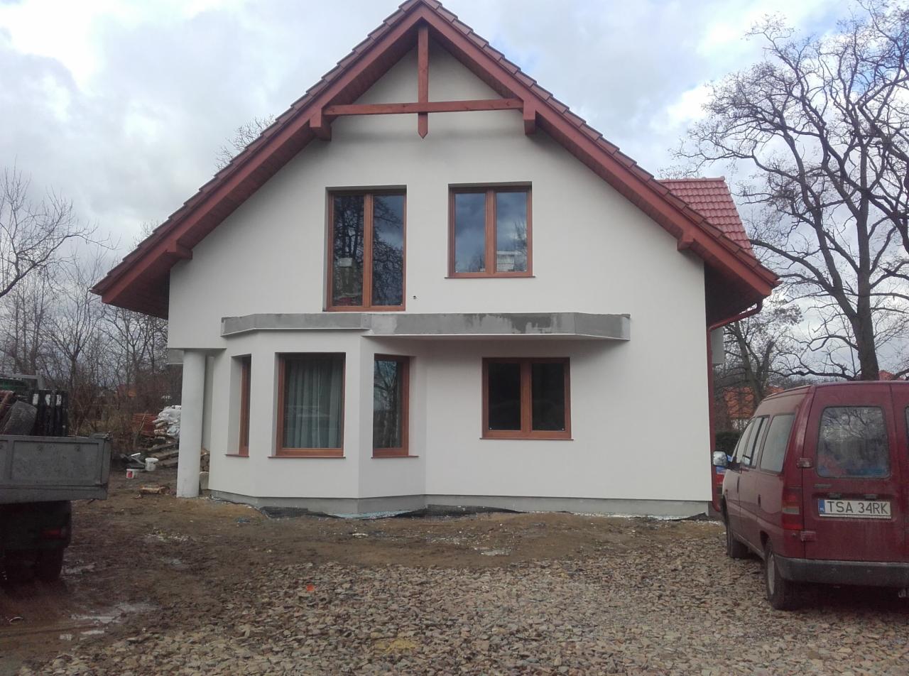 Otynkowany dom jednorodzinny z brązowymi oknami i czerwoną dachówką. Widoczny fragment samochodu i materiały budowlane. Dom po remoncie elewacji.