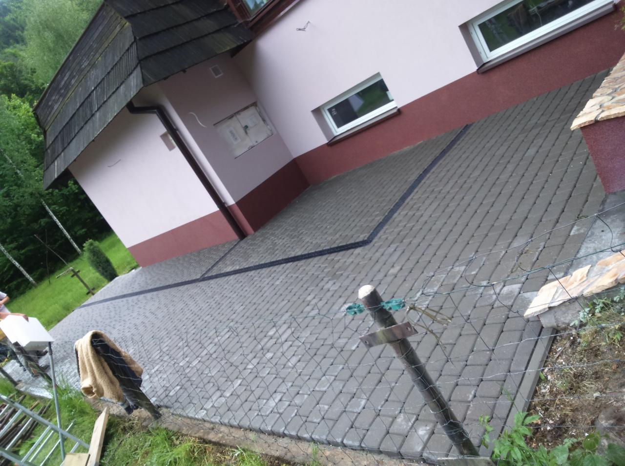 Ułożona kostka brukowa na podjeździe domu jednorodzinnego, widoczne obrzeża. Dom w kolorze różowym z brązowym pasem. W tle zieleń drzew.