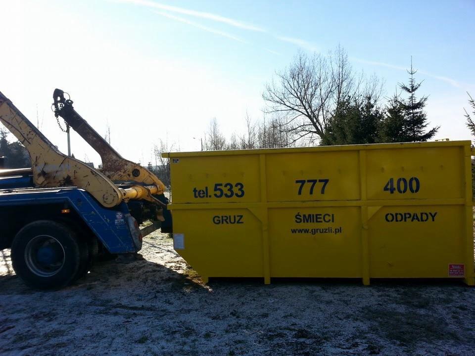 Żółty kontener na gruz i odpady, z numerami telefonu i adresem strony internetowej, umieszczony na niebieskiej ciężarówce z dźwigiem na tle drzew i błękitnego nieba.