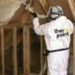 Aplikacja izolacji natryskowej pianką PUR na poddaszu, pracownik w kombinezonie ochronnym z dyszą, widoczne drewniane krokwie i fragment izolacji.
