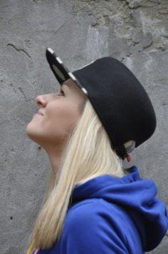 Uśmiechnięta blondynka w niebieskiej bluzie i czarnej czapce typu snapback z białymi kropkami na daszku, patrzy w górę na tle szarej, lekko popękanej ściany.