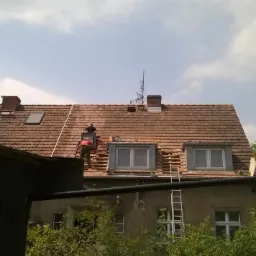 Demontaż starej dachówki na dwuspadowym dachu domu z oknami połaciowymi. Prace dekarskie w toku, widoczne elementy więźby i drabina.