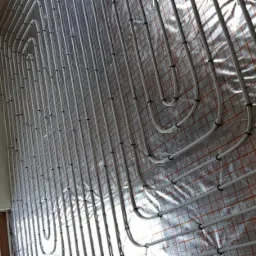 Białe rury ogrzewania podłogowego SIGMA, przymocowane czarnymi uchwytami do folii z czerwoną kratką, widok z góry, fragment ściany i okna w tle.