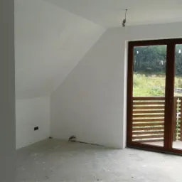 Wnętrze poddasza z oknem balkonowym i świeżo otynkowanymi na biało ścianami. Widoczna surowa posadzka i wystający kabel. Na zewnątrz widok na zieleń.