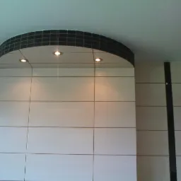 Wykończona ściana z jasnych, prostokątnych płytek ceramicznych, z półokrągłym elementem dekoracyjnym z czarnych płytek u góry i trzema wbudowanymi punktami świetlnymi.