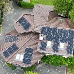 Widok z góry na dom z brązowym dachem krytym dachówką i panelami słonecznymi, otoczony zielenią i brukowanym podjazdem