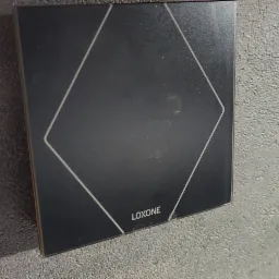 Czarny panel sterowania Loxone z białym obrysem rombu, zamontowany na szarej, teksturowanej ścianie.