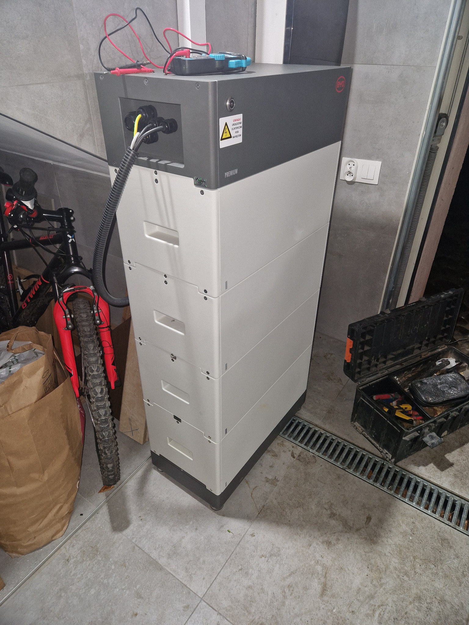 Biała, modułowa bateria energii BYD Premium LVL 15.4, podłączona przewodami, obok rower górski i skrzynka z narzędziami w garażu z szarą, kafelkową podłogą i kratką odpływową.
