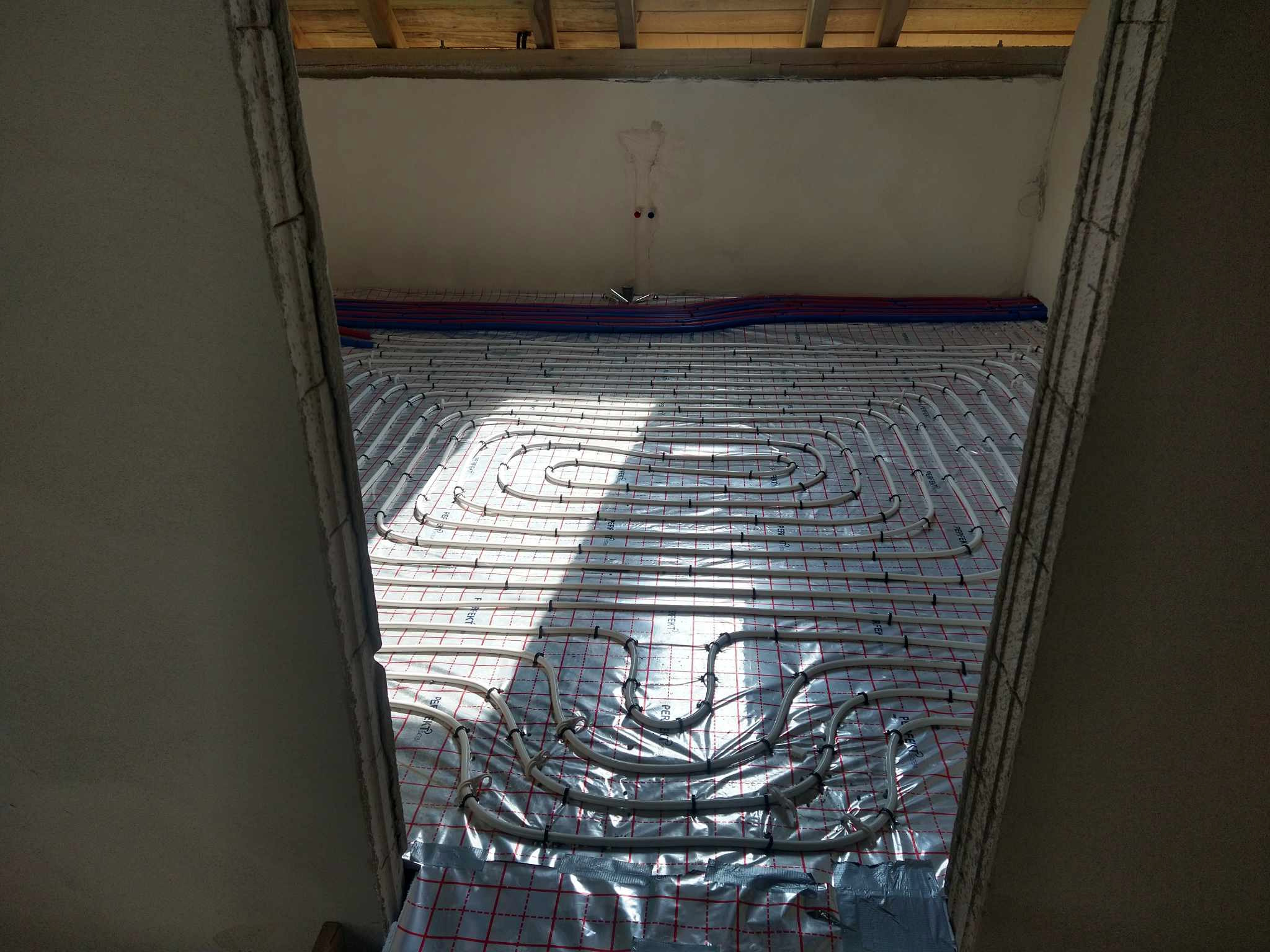 Rozłożona instalacja ogrzewania podłogowego na folii z siatką, widok z góry w surowym stanie, z widocznymi rurkami i elementami mocującymi, w tle sufit z wystającymi przewodami.