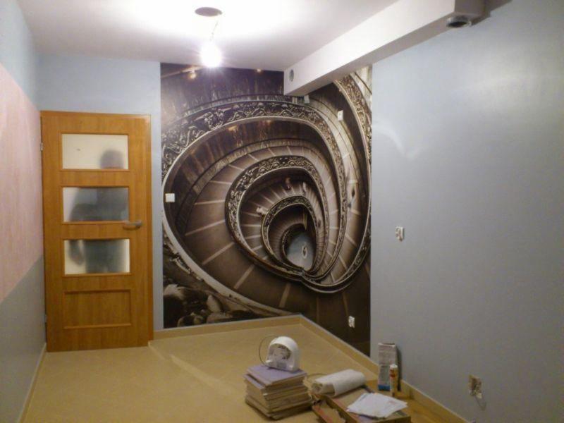 Pokój po tapetowaniu: fototapeta ze spiralnymi schodami, drewniane drzwi z szybami, jasne ściany i podłoga. Widoczne narzędzia i materiały.