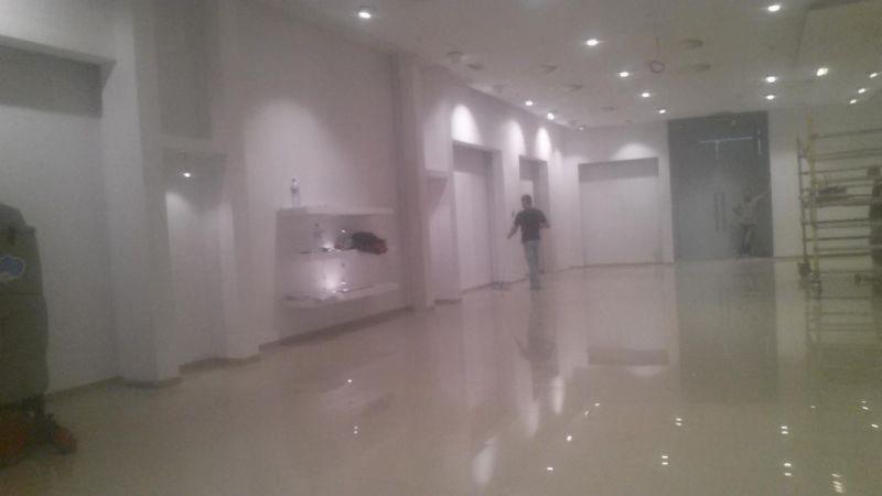 Błyszcząca, jasna posadzka w przestronnym wnętrzu z białymi ścianami i oświetleniem punktowym. Widoczny fragment regału z ekspozycją i osoba w tle.