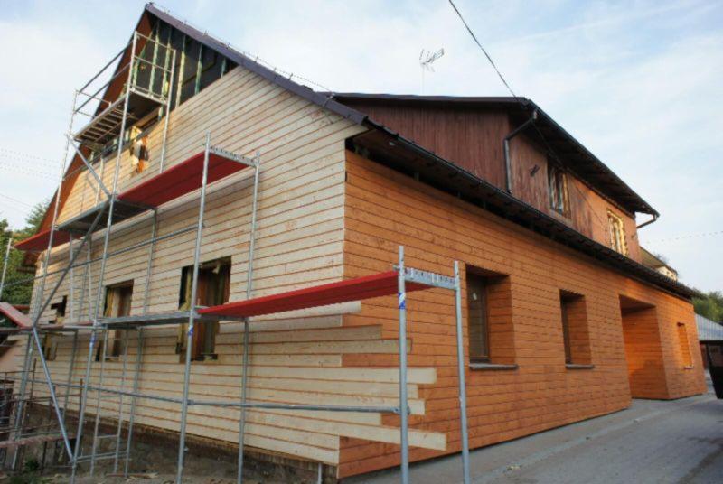 Dom w trakcie prac wykończeniowych elewacji drewnianej. Część ściany w surowym drewnie, druga część pomalowana na brązowo. Widoczne rusztowania.