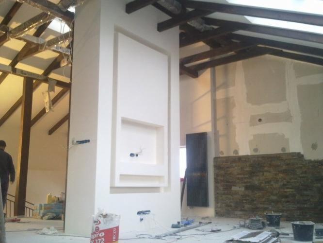 Remont wnętrza w stylu loft: biała ściana z dekoracyjnym wnęką, surowa drewniana konstrukcja dachu, ściana z kamienia dekoracyjnego i grzejnik. Widoczny worek z gładzią.