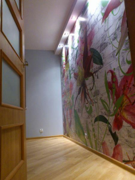 Pokój po remoncie: drewniana podłoga, ściana z tapetą w kwiaty na tle cegieł, oświetlenie sufitowe. Widok zza otwartych drzwi.