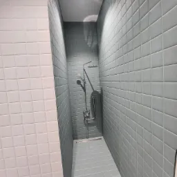 Wąska kabina prysznicowa wyłożona błękitnymi i białymi kwadratowymi płytkami, z chromowaną armaturą i odpływem liniowym w podłodze.