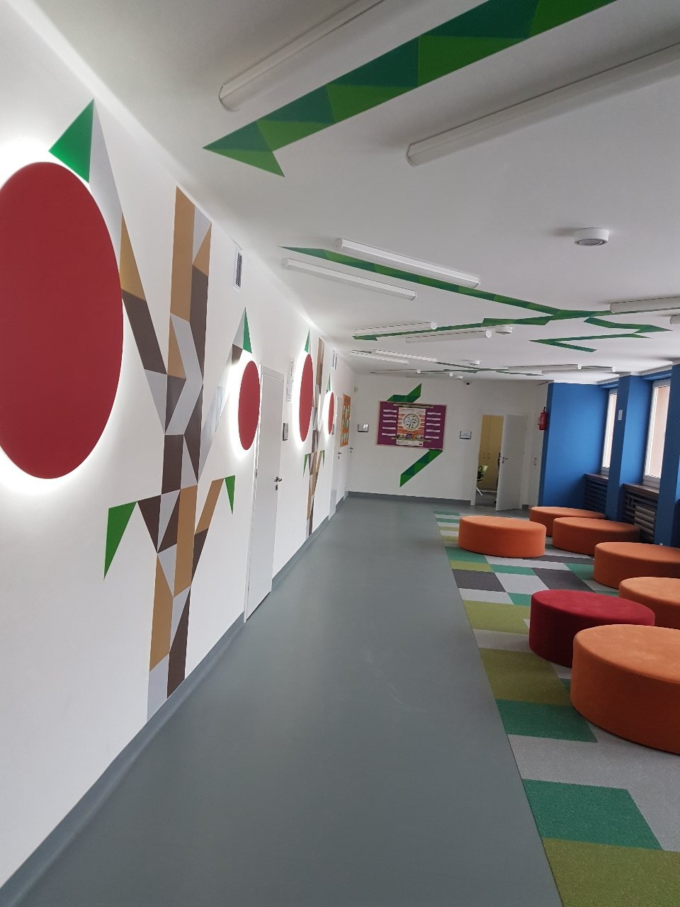 Korytarz biurowy z geometrycznymi malowidłami na ścianach, szarą podłogą i kolorowymi pufami do siedzenia, oświetlony lampami sufitowymi.