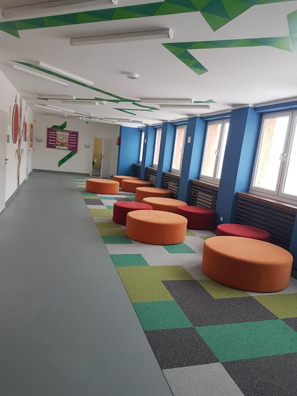 Długi korytarz w biurze lub szkole z niebieskimi ścianami, rzędem okien, pomarańczowymi pufami i wzorzystą wykładziną podłogową w odcieniach zieleni i szarości, z geometrycznymi dekoracjami...