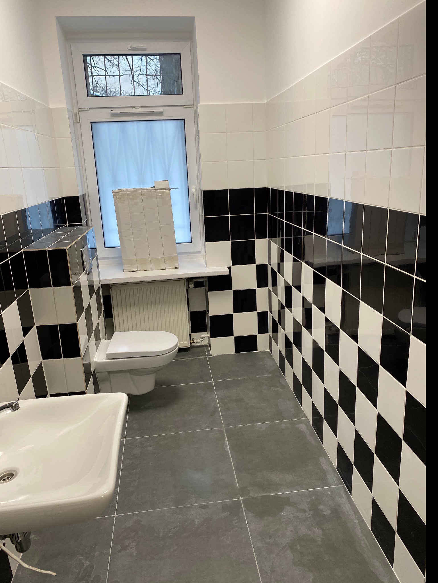 Ukończona łazienka z czarno-białymi płytkami ułożonymi w szachownicę na ścianach, szarą podłogą i białą armaturą, z paczką płytek opartą o okno.