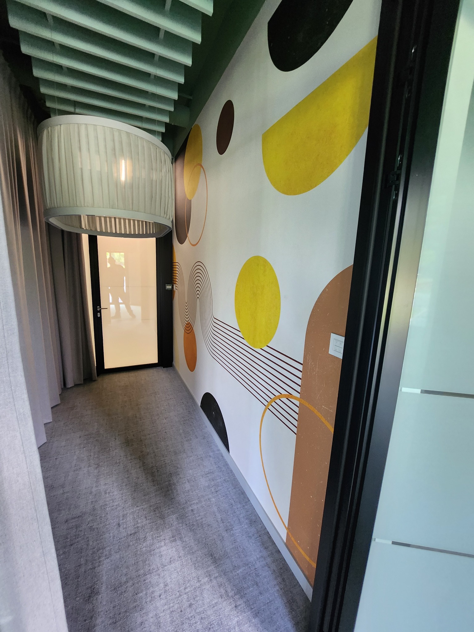 Nowoczesny korytarz z szarą wykładziną, abstrakcyjną tapetą w geometryczne wzory w odcieniach żółci i brązu, oraz wiszącą lampą z abażurem z delikatnej tkaniny.