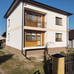 Dwupiętrowy dom z białą elewacją, drewnianymi akcentami i czarnymi rynnami, sfotografowany w słoneczny dzień z perspektywy lekko od dołu, z widocznym fragmentem ogrodzenia z żółtej cegły.